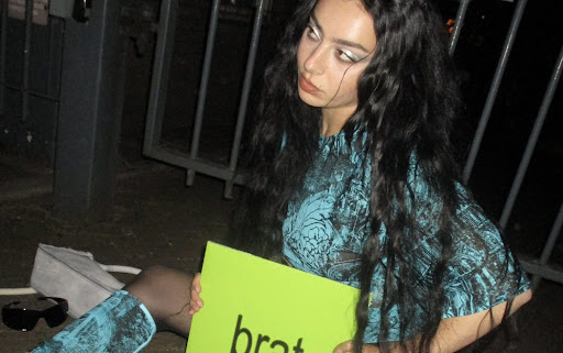 Photo d'une fille assise au sol tenant une pancart qui affiche "brat"